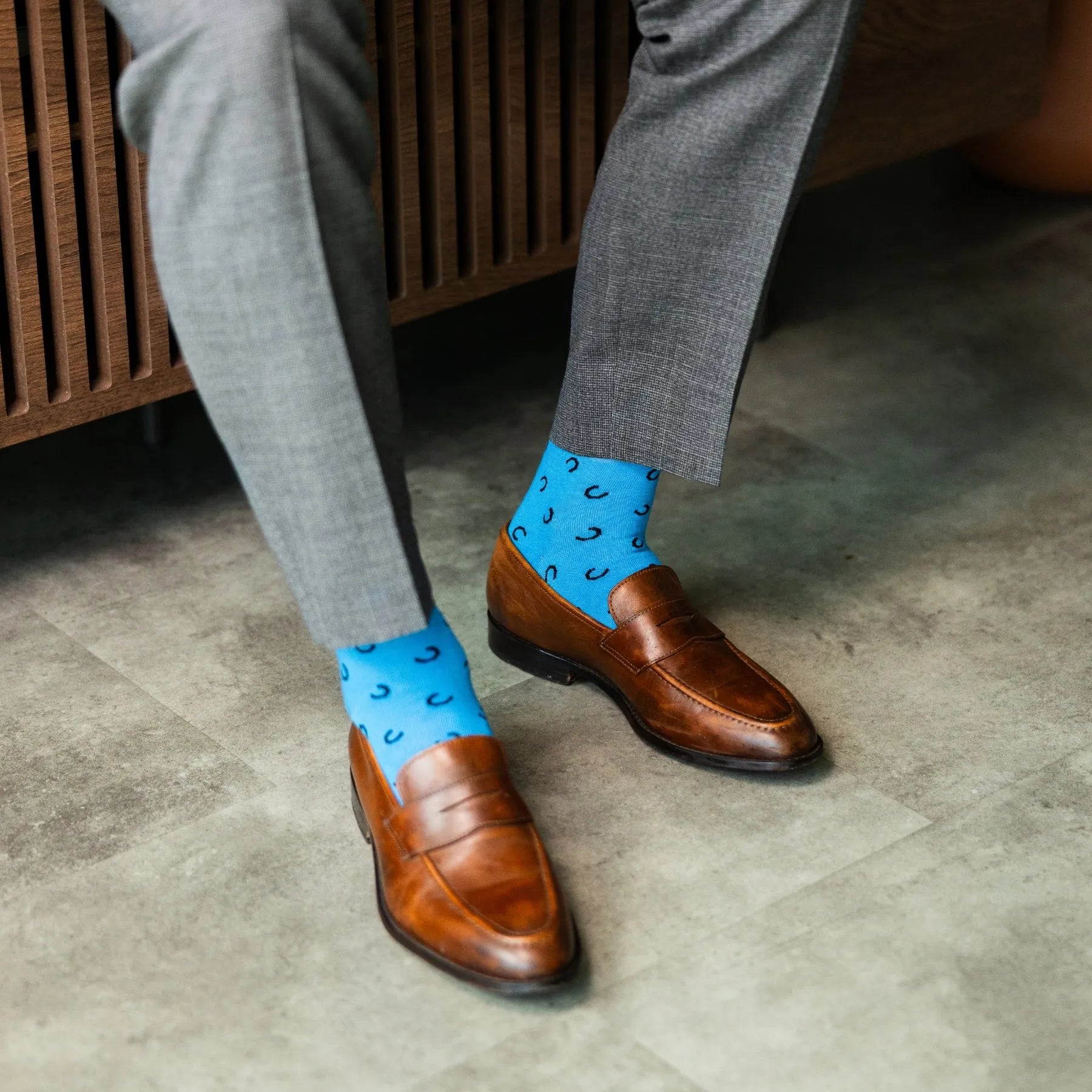 Person wearing gray pants and brown shoes with blue socks featuring black patterns on a concrete floor.