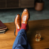 Person wearing brown leather shoes and red patterned socks with a glass of whiskey on a wooden table.