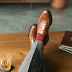 Person wearing brown leather shoes and red patterned socks, with a glass of whiskey on a wooden table.