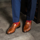 Brown loafers with red patterned socks worn with blue pants on a gray floor.