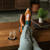 Brown leather shoes with green socks on a wooden table, accompanied by a glass of whiskey.