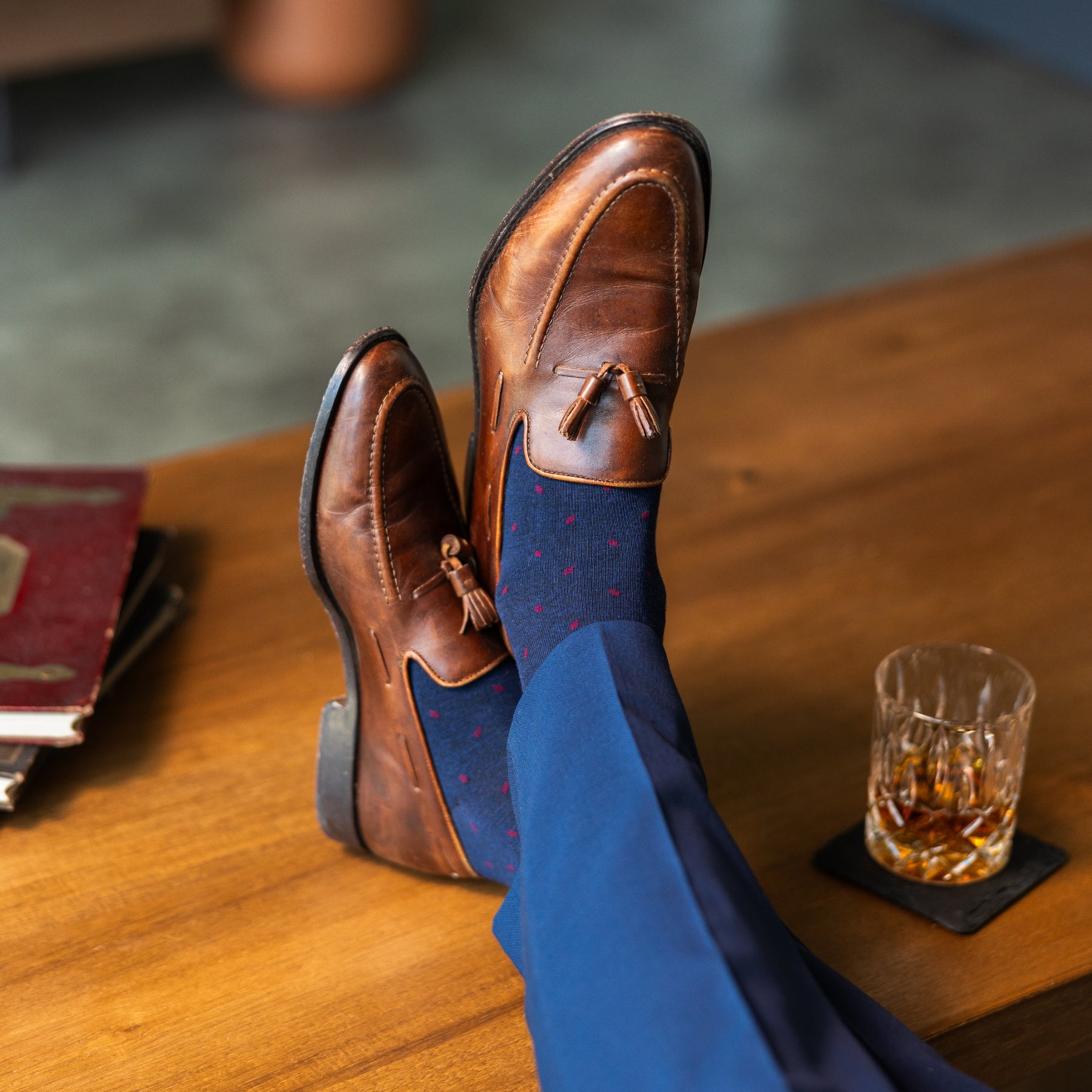 Navy blue socks with a red pattern worn with navy trousers and brown tassel loafers, sitting on a wooden table with a glass of whiskey.