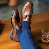 Navy blue socks with a red pattern worn with navy trousers and brown tassel loafers, sitting on a wooden table with a glass of whiskey.