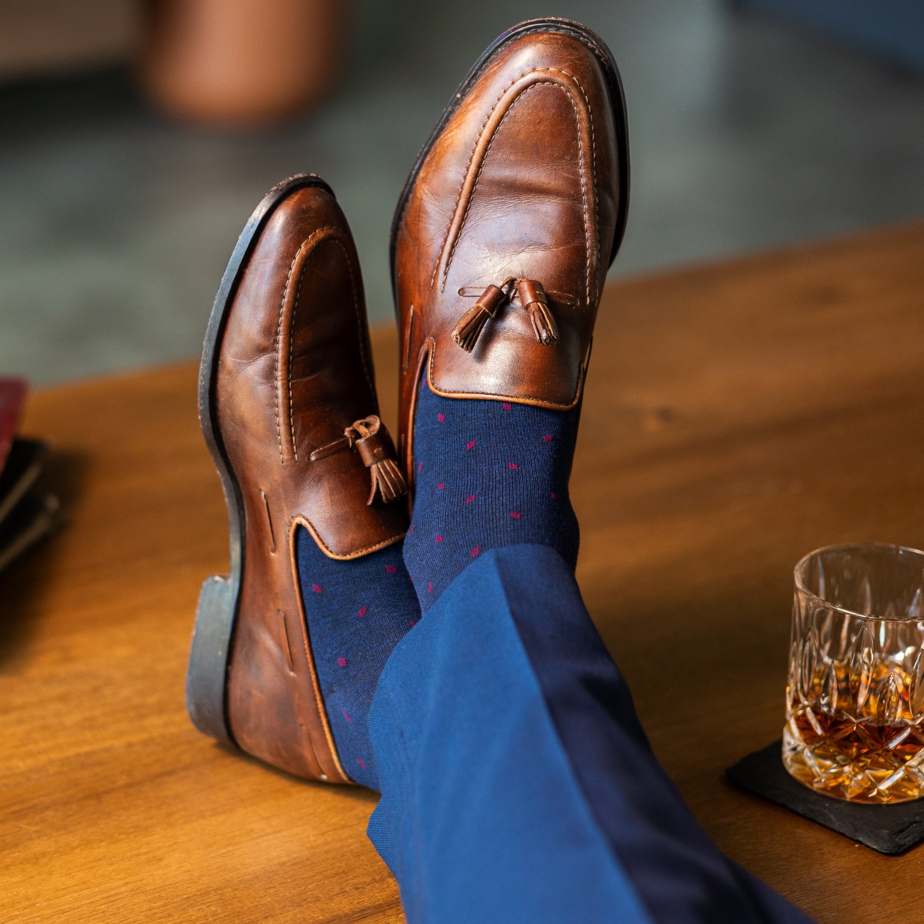 Navy blue socks with a red pattern worn with navy trousers and brown tassel loafers, sitting on a wooden table with a glass of whiskey.