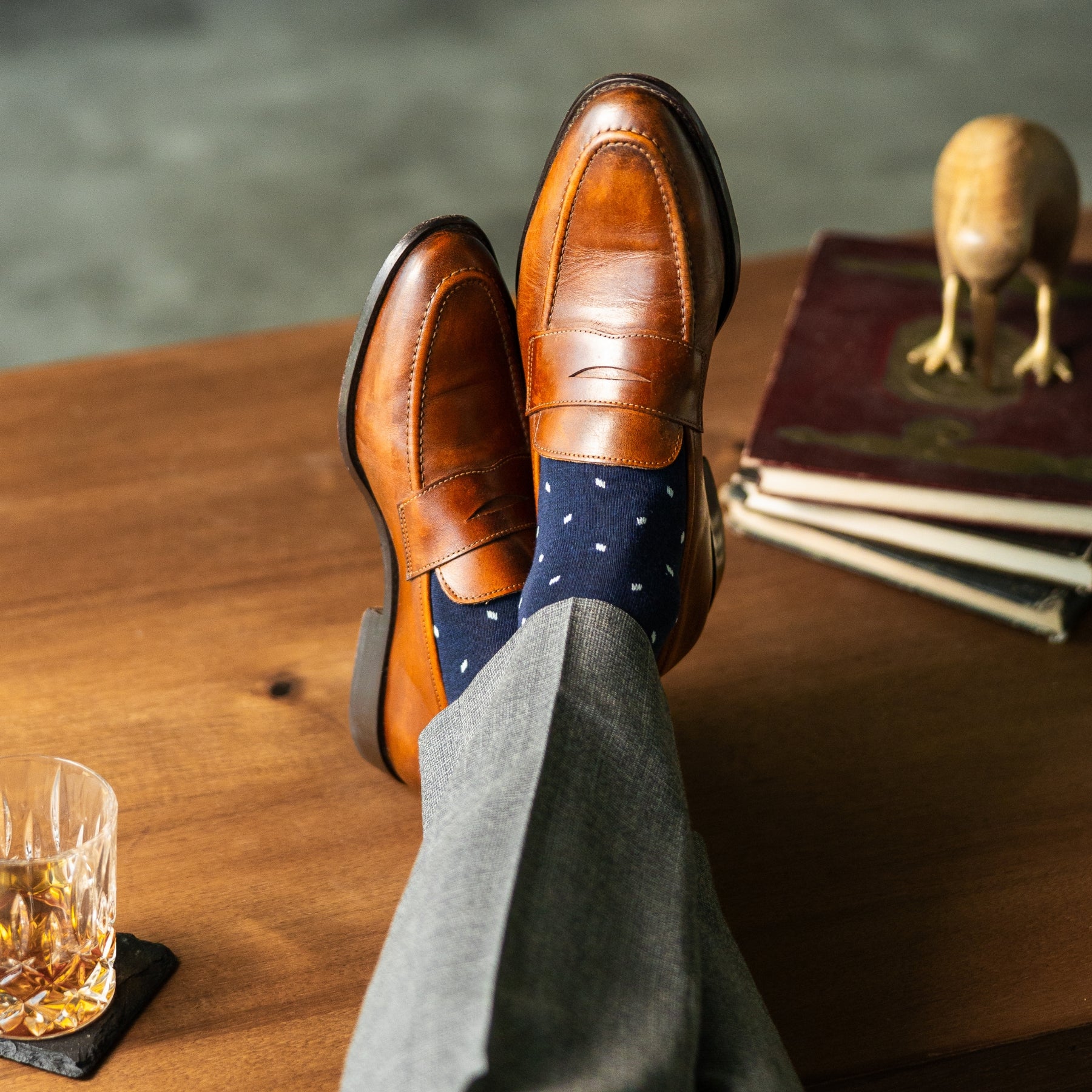 Brown leather shoes, navy polka dot socks, and gray pants on a wooden table with a glass of whiskey.