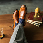 Brown leather shoes with blue polka dot socks on a wooden table with books and a glass.