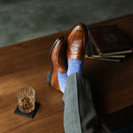 Brown leather shoes with blue patterned socks on a wooden table with a glass of whiskey.