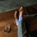 Brown leather shoes with blue patterned socks on a wooden table with a glass of whiskey.
