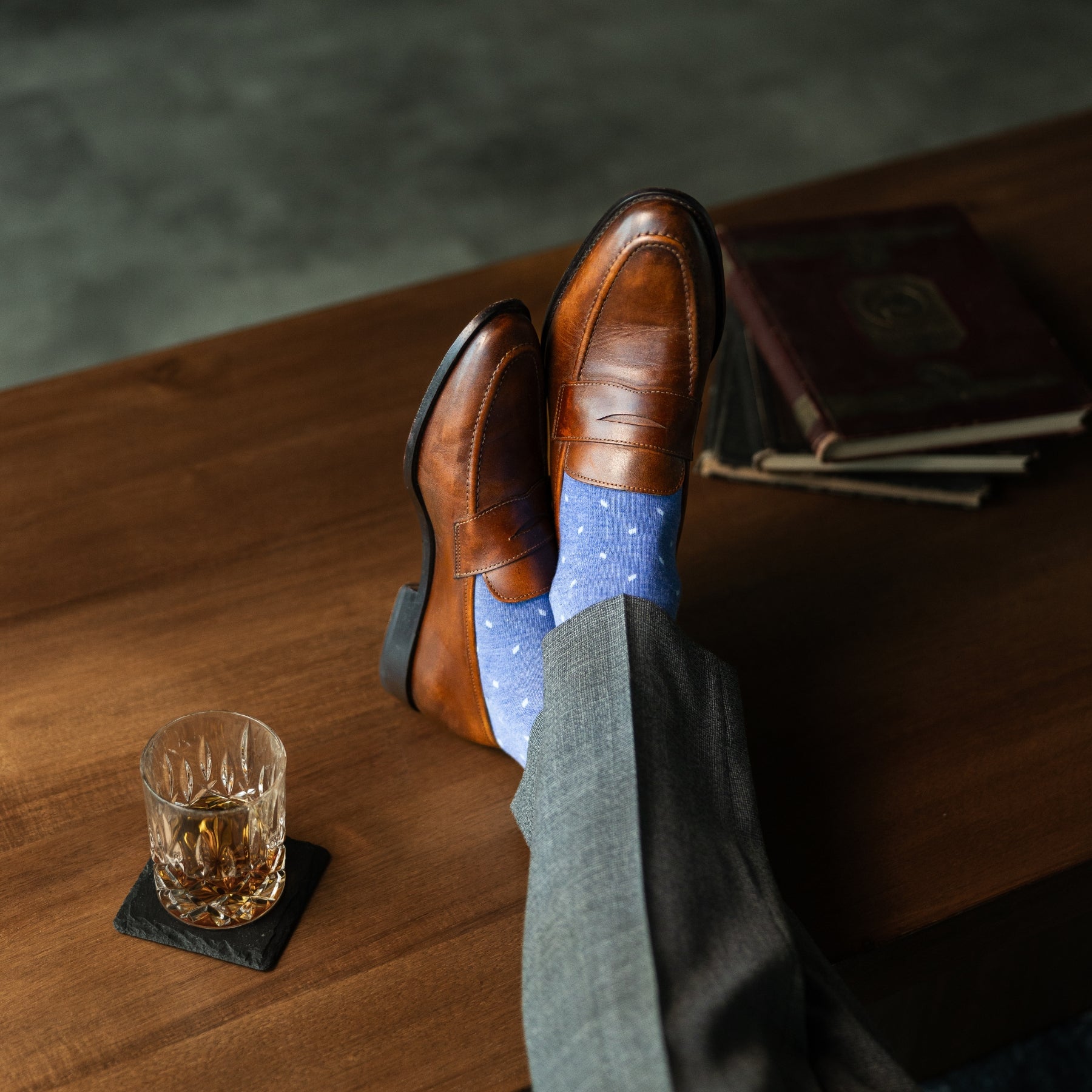 Brown leather shoes with blue patterned socks on a wooden table with a glass of whiskey.