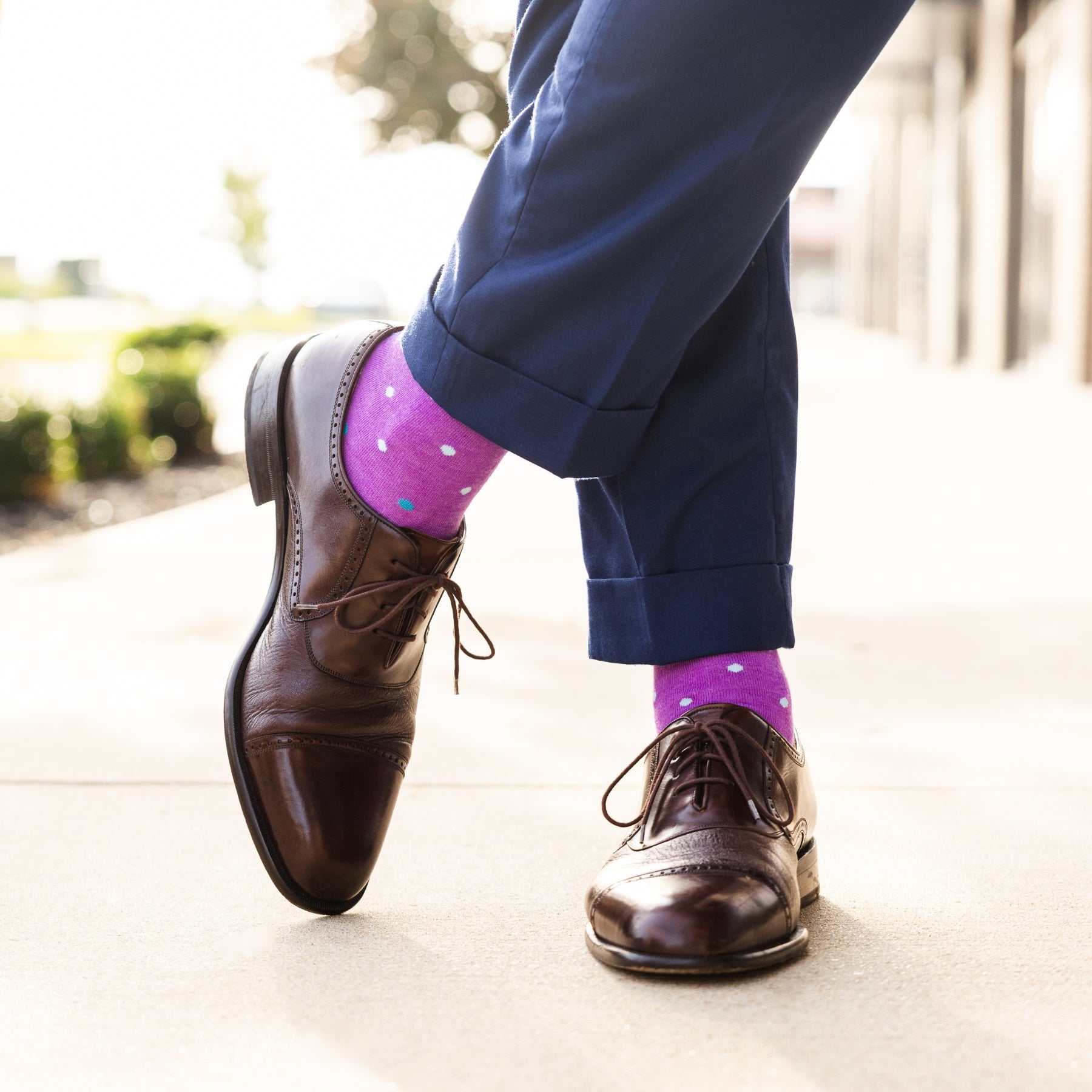 Person wearing brown dress shoes and purple socks with multi-colored polka dots on a blurred outdoor background.