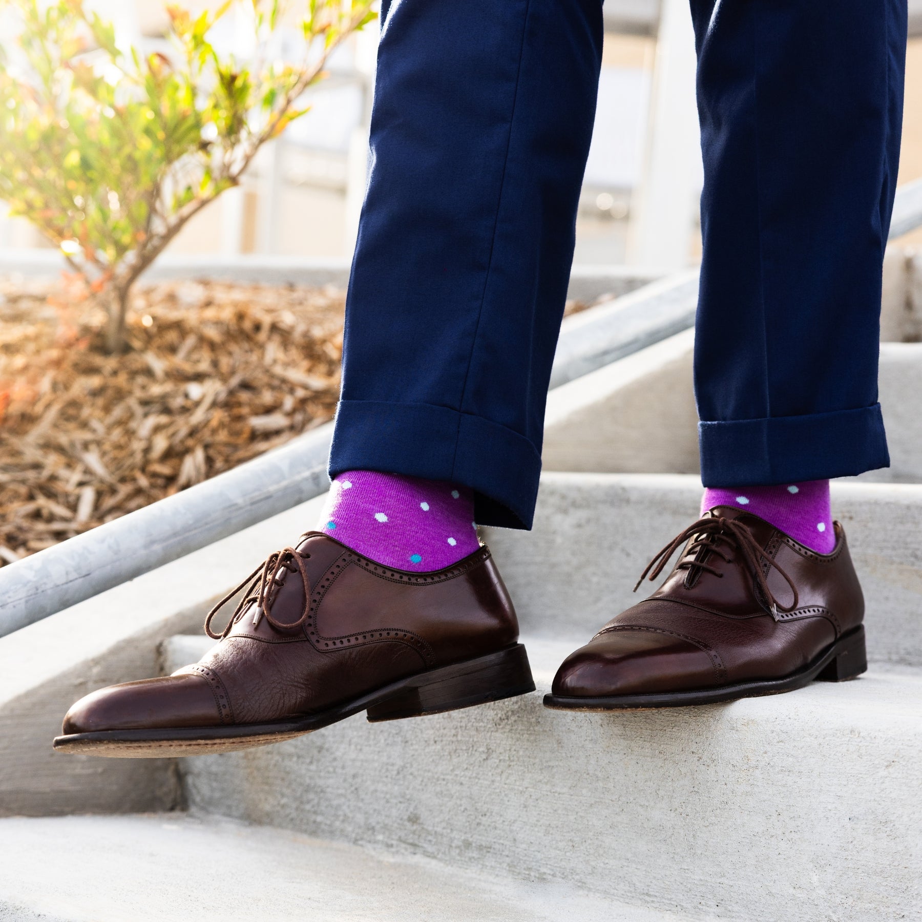 Person wearing navy trousers, brown shoes and purple socks with white polka dots on a concrete stairs.