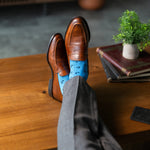 Brown leather shoes with blue socks on a wooden table, with books and a plant in the background.