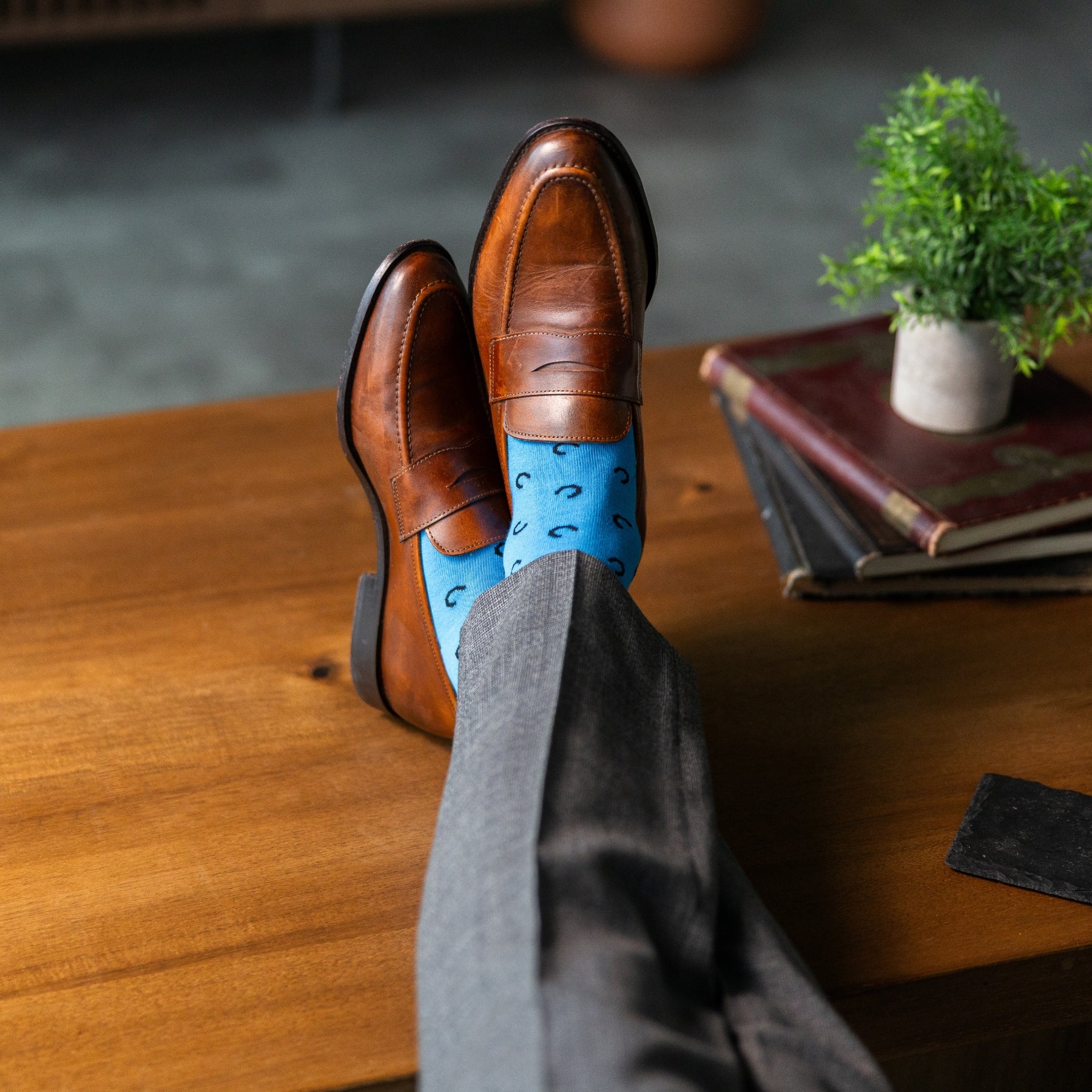Brown leather shoes with blue socks on a wooden table, with books and a plant in the background.