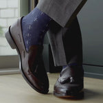 Brown leather loafers with tassels worn with blue patterned socks and gray pants on a wooden floor.