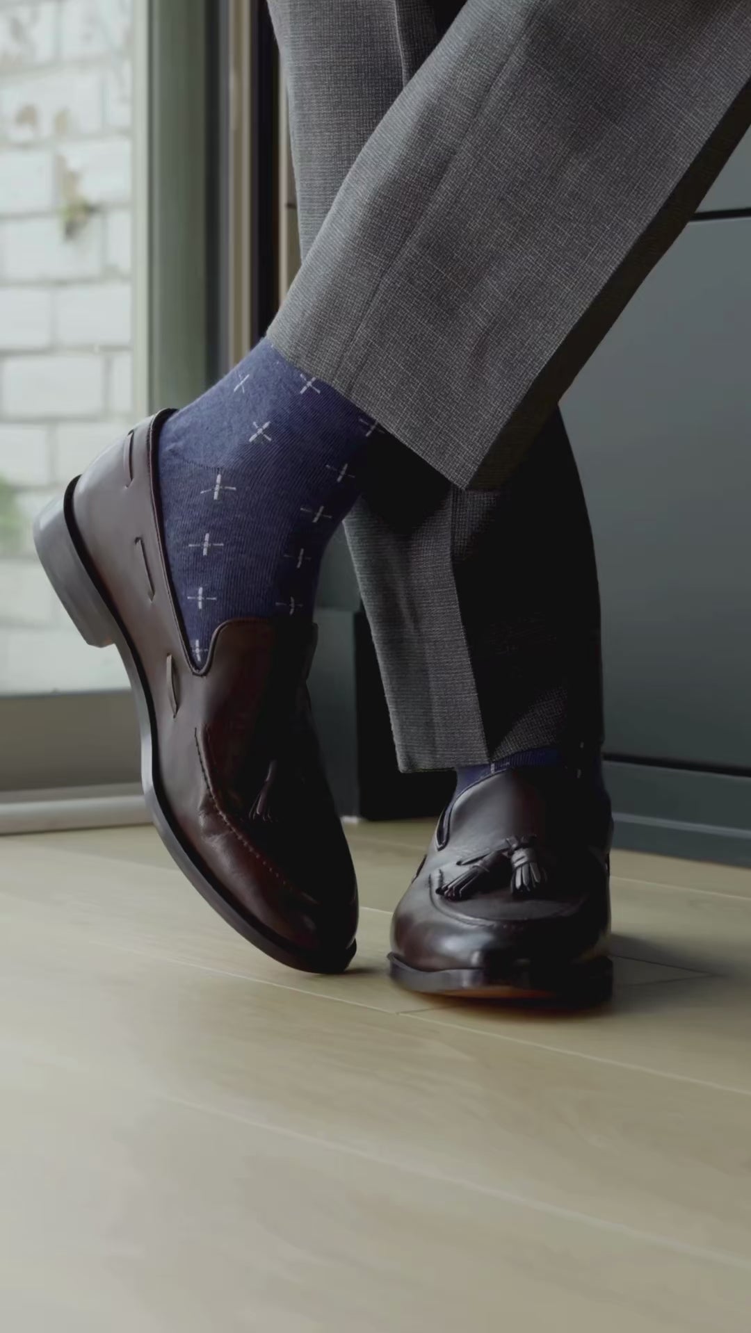 Brown leather loafers with tassels worn with blue patterned socks and gray pants on a wooden floor.