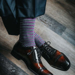 Man sitting with legs crossed wearing purple, grey, and white striped dress socks and brown dress shoes
