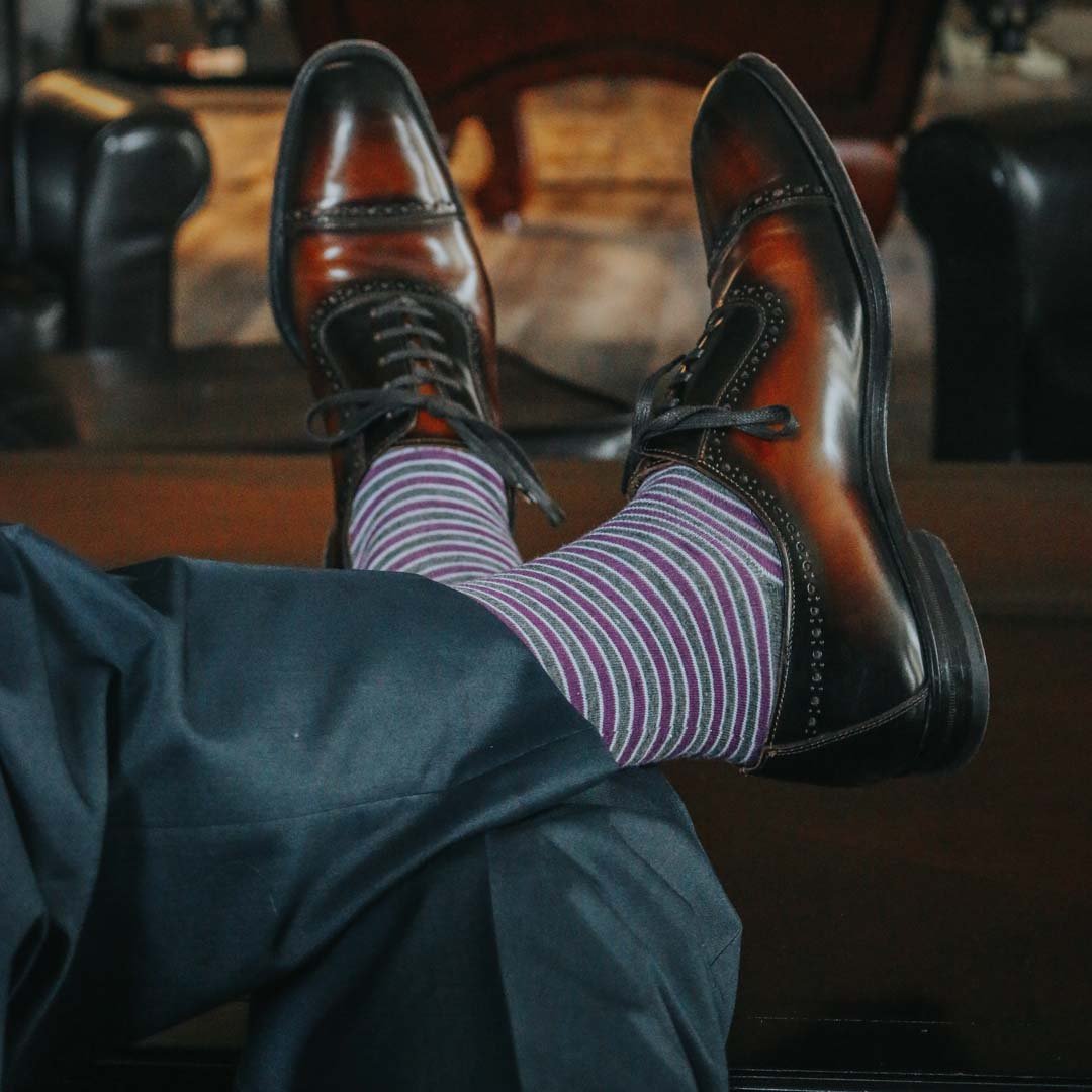 Man sitting with legs crossed wearing purple, grey, and white striped dress socks and brown dress shoes
