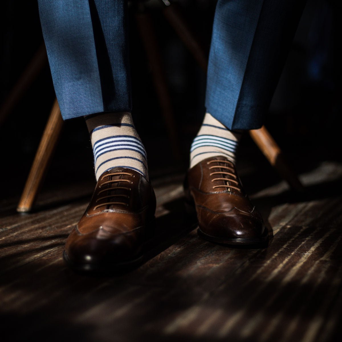 Man sitting and wearing blue trousers with tan, navy blue, and white striped dress socks and dark brown dress shoes